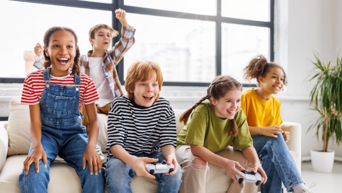 A group of children sitting on a couch, with two holding game controllers and others smiling in a bright room.