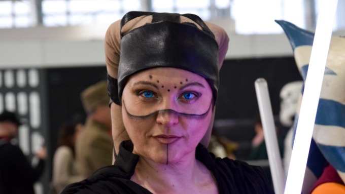 A woman in black cosplay makeup and headwear holding a glowing purple sword at an indoor convention.