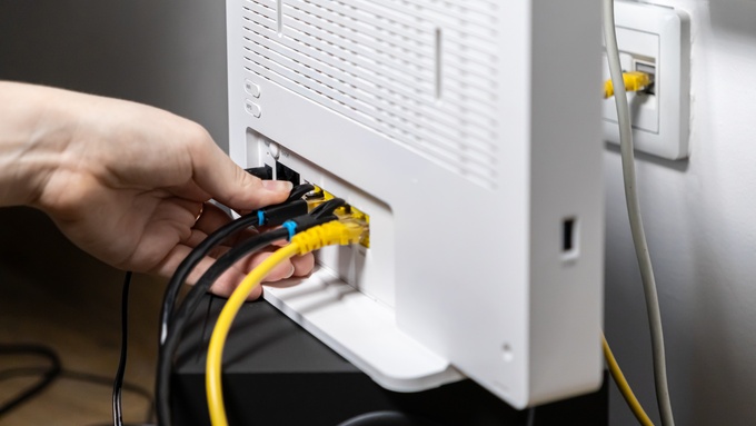 A close-up view shows a person plugging a black cable into a white Wi-Fi router. There are other cables plugged in, too.